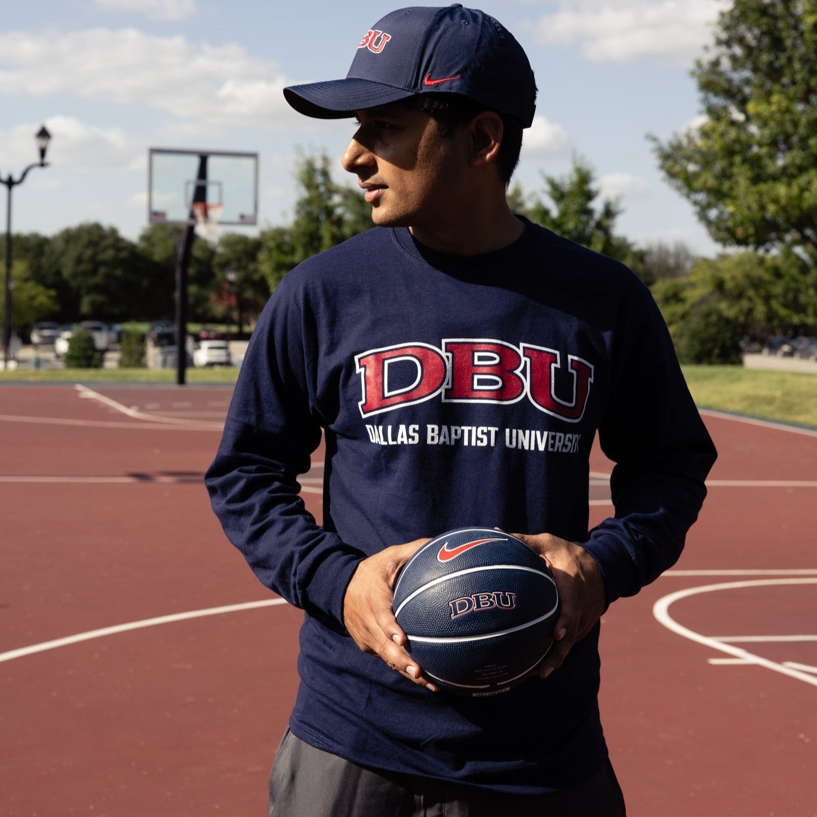 Person wearing a navy long-sleeve shirt and cap with 'DBU' holding a basketball on a court.