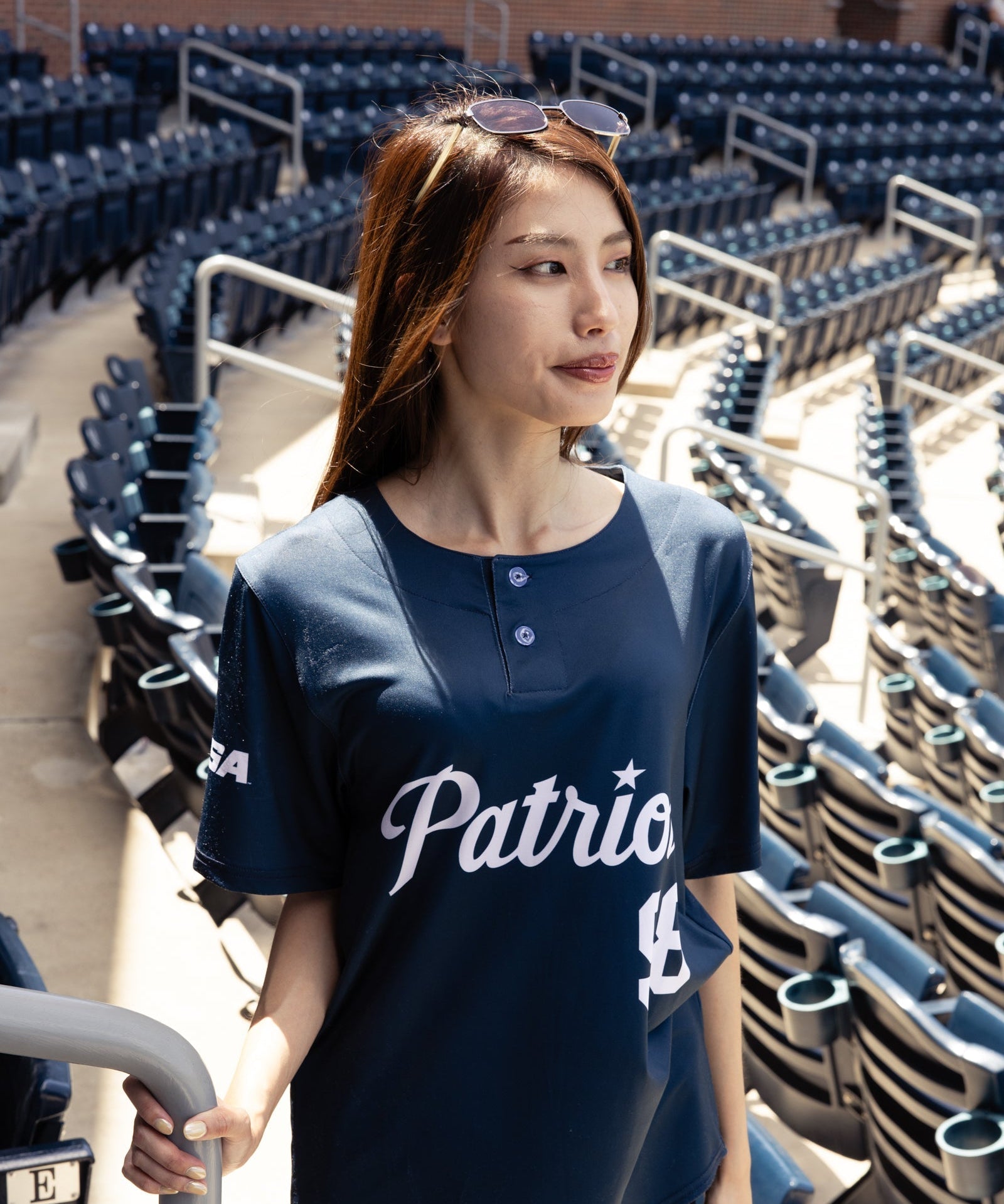 Woman wearing a navy blue 'Patriots' shirt in an empty stadium.